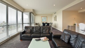 A modern living room with a leather sofa, a coffee table, and a dining area with chairs and a table. - The Waterfront Apartments, Geelong (Geelong)