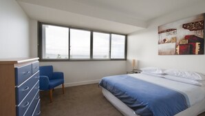 A bedroom with a bed, a desk with drawers, a chair, a window, and a wall with artwork. - The Waterfront Apartments, Geelong (Geelong)