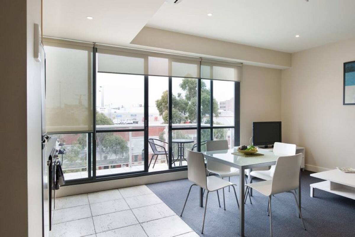 Un espacio de oficina moderno con un ventanal grande, una mesa de comedor con sillas, un escritorio blanco y un cuadro en la pared.