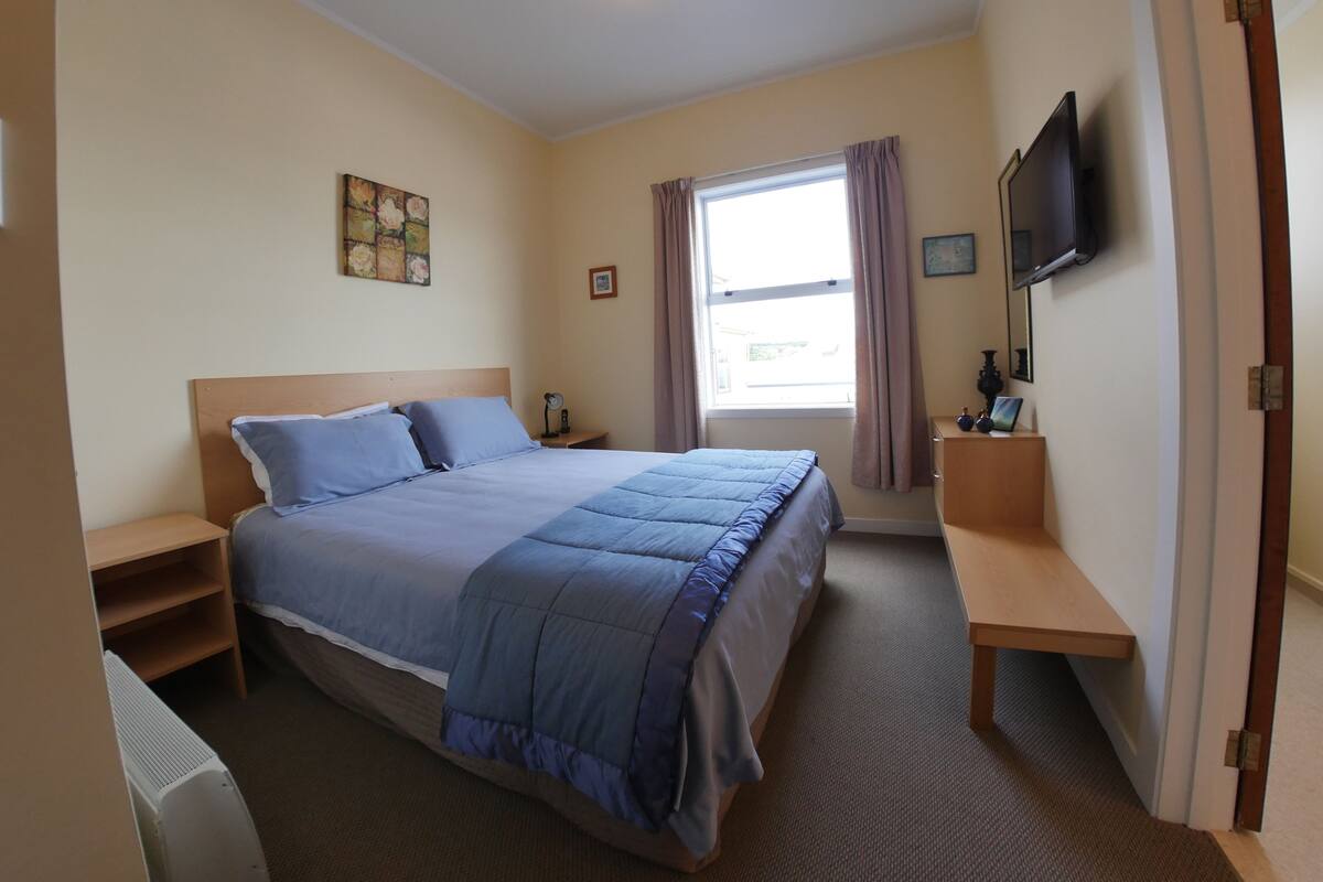 Habitación de hotel con una cama grande, una mesita de noche, un televisor montado en la pared y una ventana con cortinas.