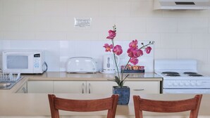 A kitchen with white appliances, a microwave, toaster, kettle, and stove. There is a wooden table with chairs and a potted orchid plant. - JE Resort (Avonside)