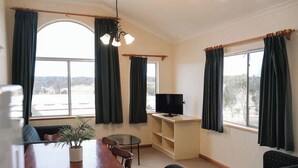 A living room with a wooden dining table, a glass-top coffee table, a sofa, a TV on a stand, and large windows with curtains. - JE Resort (Avonside)