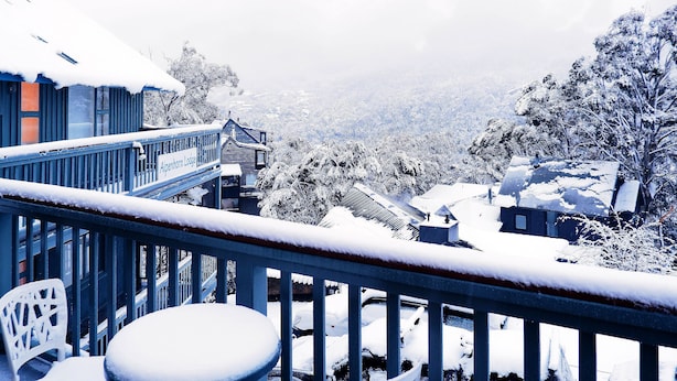 A snowy balcony with a chair, a sign reading "Alpenhorn Lodge," and a view of snow-covered buildings and trees. - Alpenhorn (Thredbo)