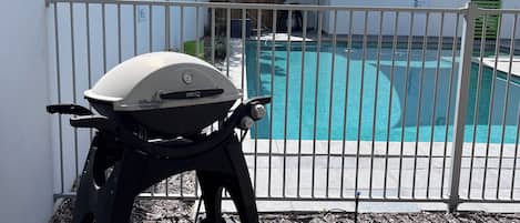 A barbecue grill near a swimming pool, enclosed by a white fence with a metal gate.