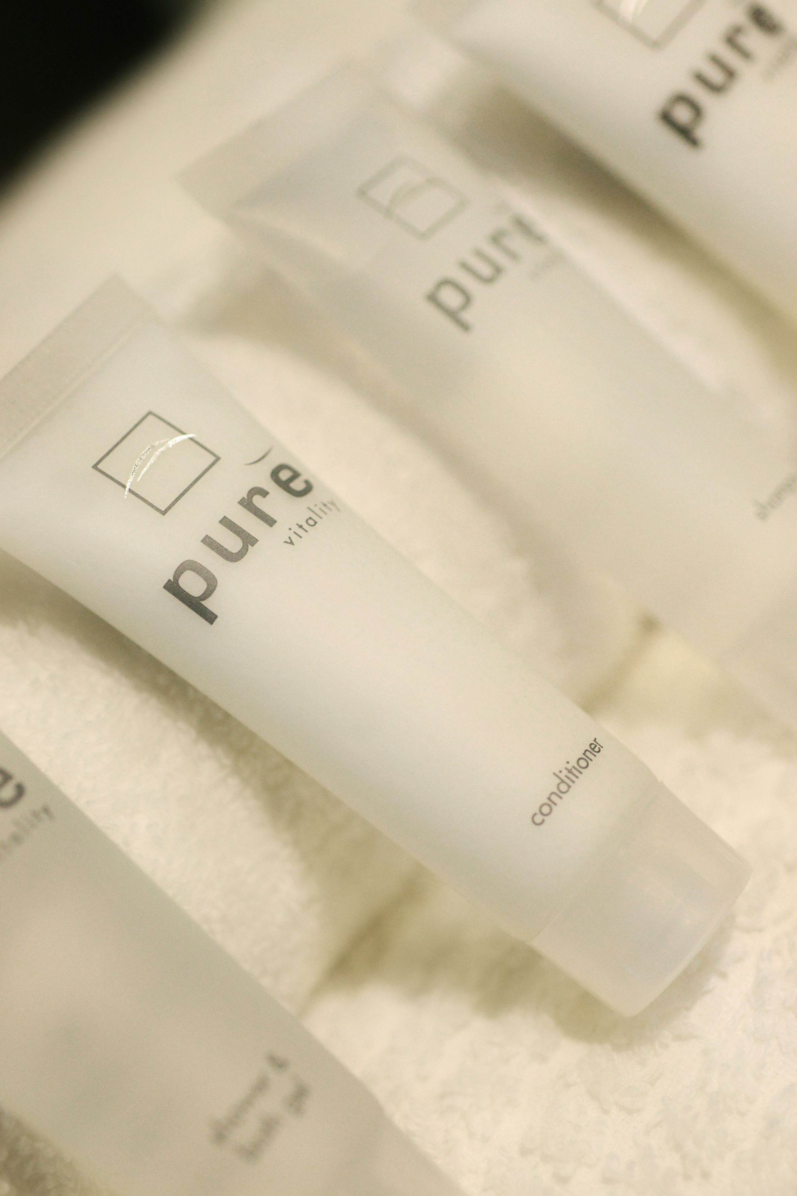 Primer plano de varios tubos blancos etiquetados como 'pure vitality conditioner' sobre una toalla blanca.