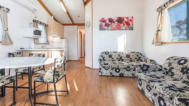Ein gemütliches Wohnzimmer mit einer Couch und Sesseln, einem Esstisch mit Stühlen, einer Küchenzeile mit Mikrowelle und Herd sowie einem großen Fenster mit Vorhängen.