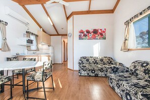 En hyggelig stue med sofa og lænestole, et spisebord med stole, et køkkenområde med mikroovn og komfur, samt et stort vindue med gardiner.