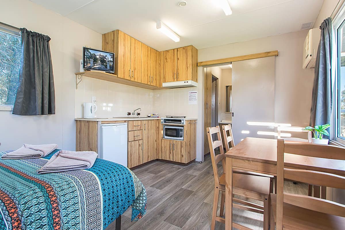A compact room with a bed, a small kitchenette, a dining table with chairs, and a window with curtains.