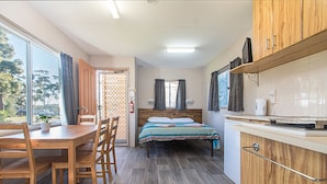 Ein gemütlicher Raum mit Esstisch aus Holz, Stühlen, einem Bett mit gemustertem Überwurf, einer kleinen Küchenzeile und einem großen Fenster mit Vorhängen.