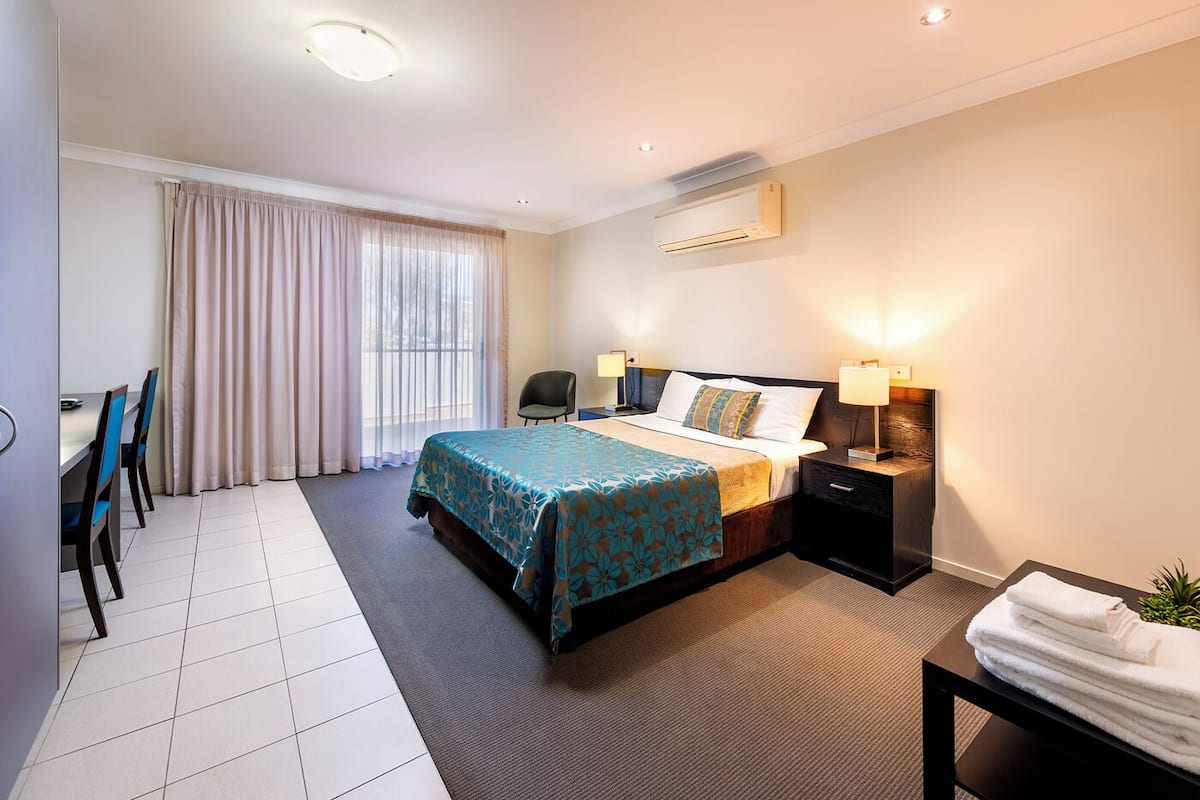 Habitación de hotel con cama, mesitas de noche, escritorio con sillas, zona de comedor y ventana con cortinas.