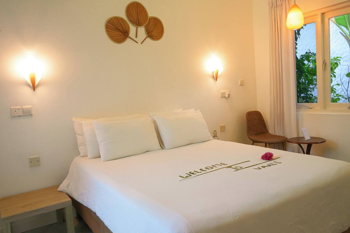 A neatly made bed with a welcome mat and a flower decoration, two wall-mounted lights, a small table, and a chair in a room with a window.