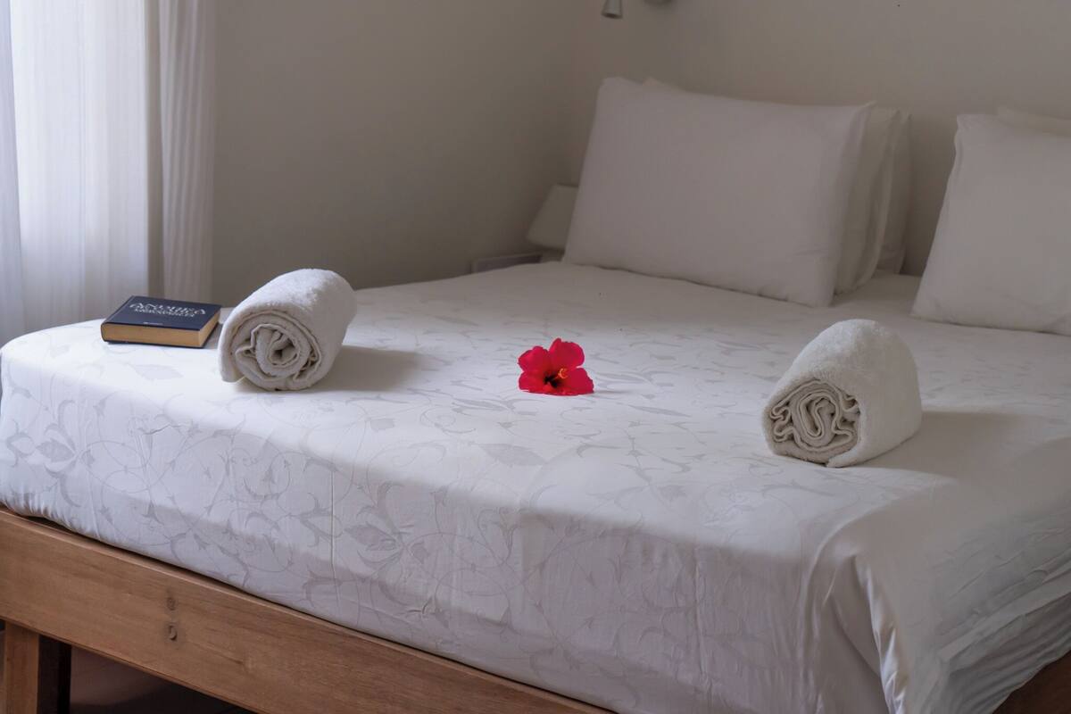 A neatly made bed with a floral-patterned bedspread, a rolled towel, a red flower, and a book on the left side.