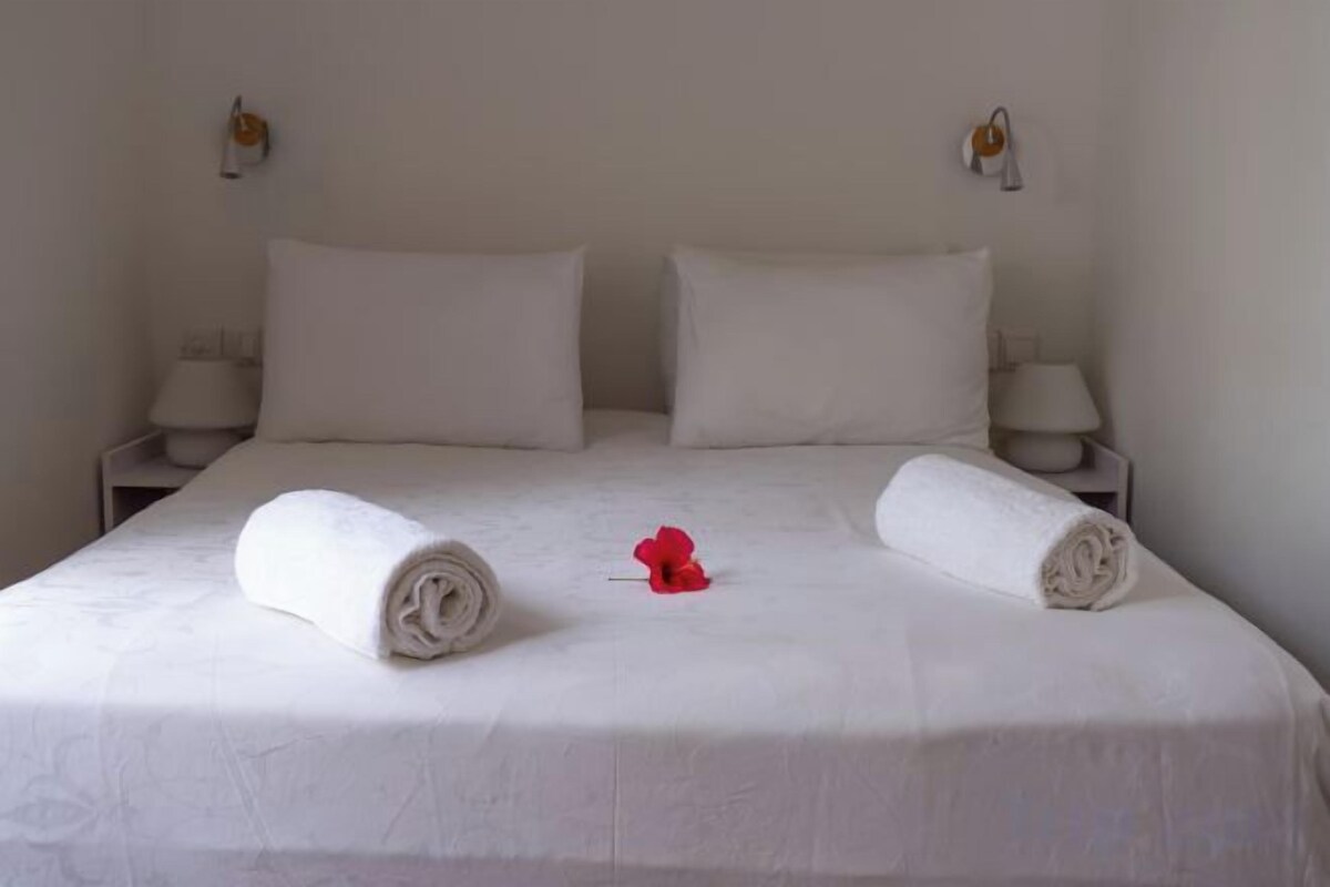 Una cama bien hecha con dos toallas enrolladas y una flor roja sobre el edredón.