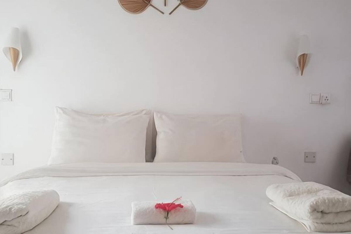 A neatly made bed with white linens, a folded towel with a red flower, and a wall-mounted light fixture.
