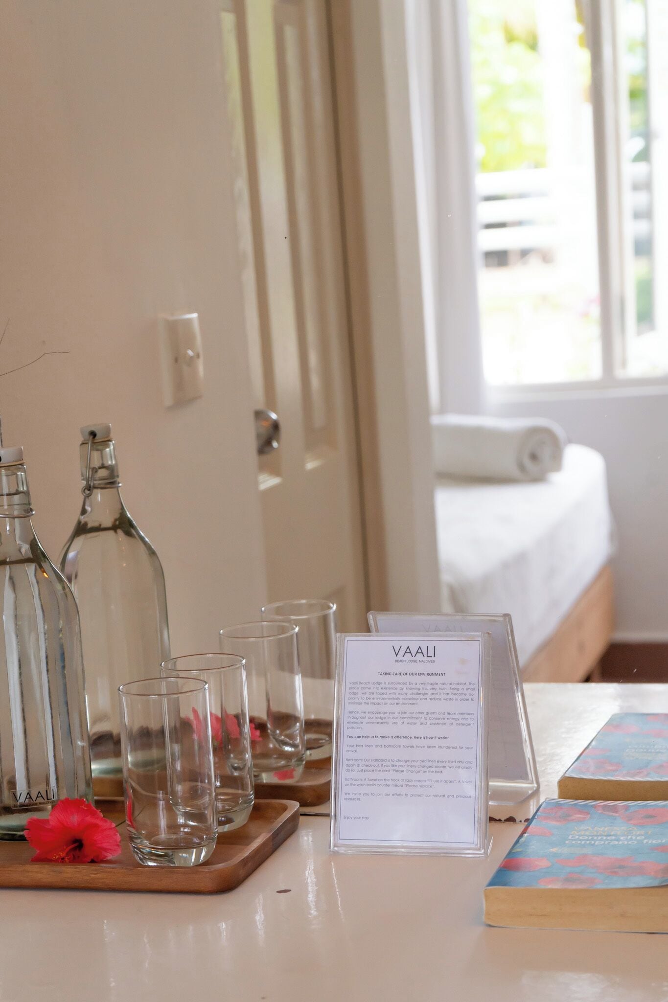 A hotel room with a bed, a bedside table with a tray holding glasses and bottles, and a sign with text.