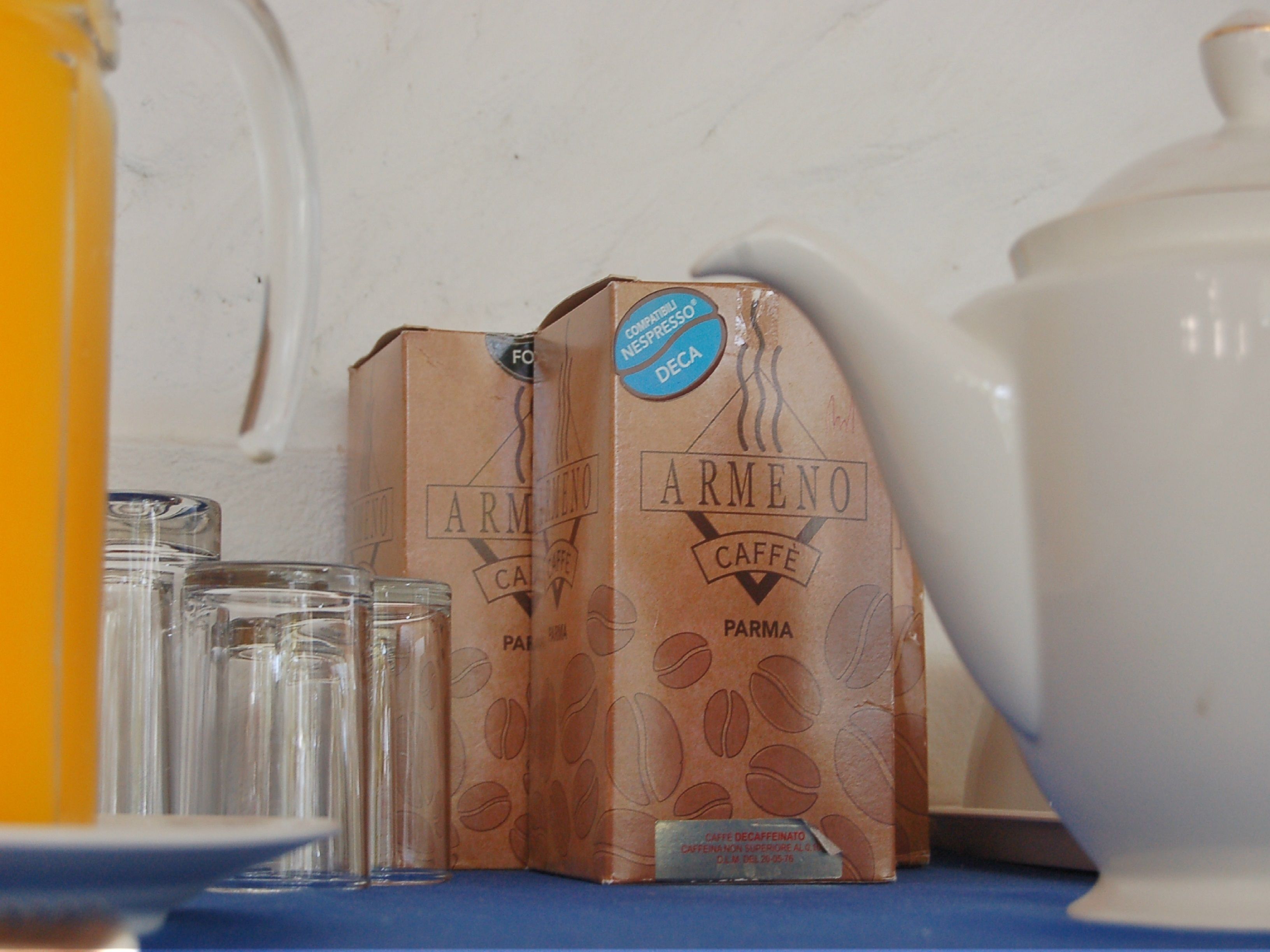Two cardboard boxes labeled 'ARMENO Caffè' with a coffee logo, placed on a blue surface next to a white teapot and clear glasses.