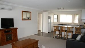 A living room with a TV, wooden furniture, and a kitchen in the background. - Breakaway Apartments (Cowra)