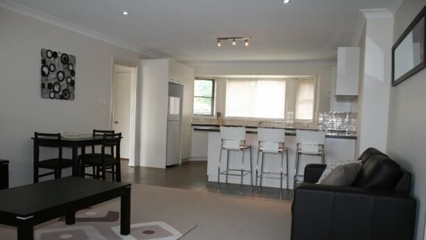 A modern living room with a black sofa, a dining table with chairs, and a kitchen area in the background. - Breakaway Apartments (Cowra)