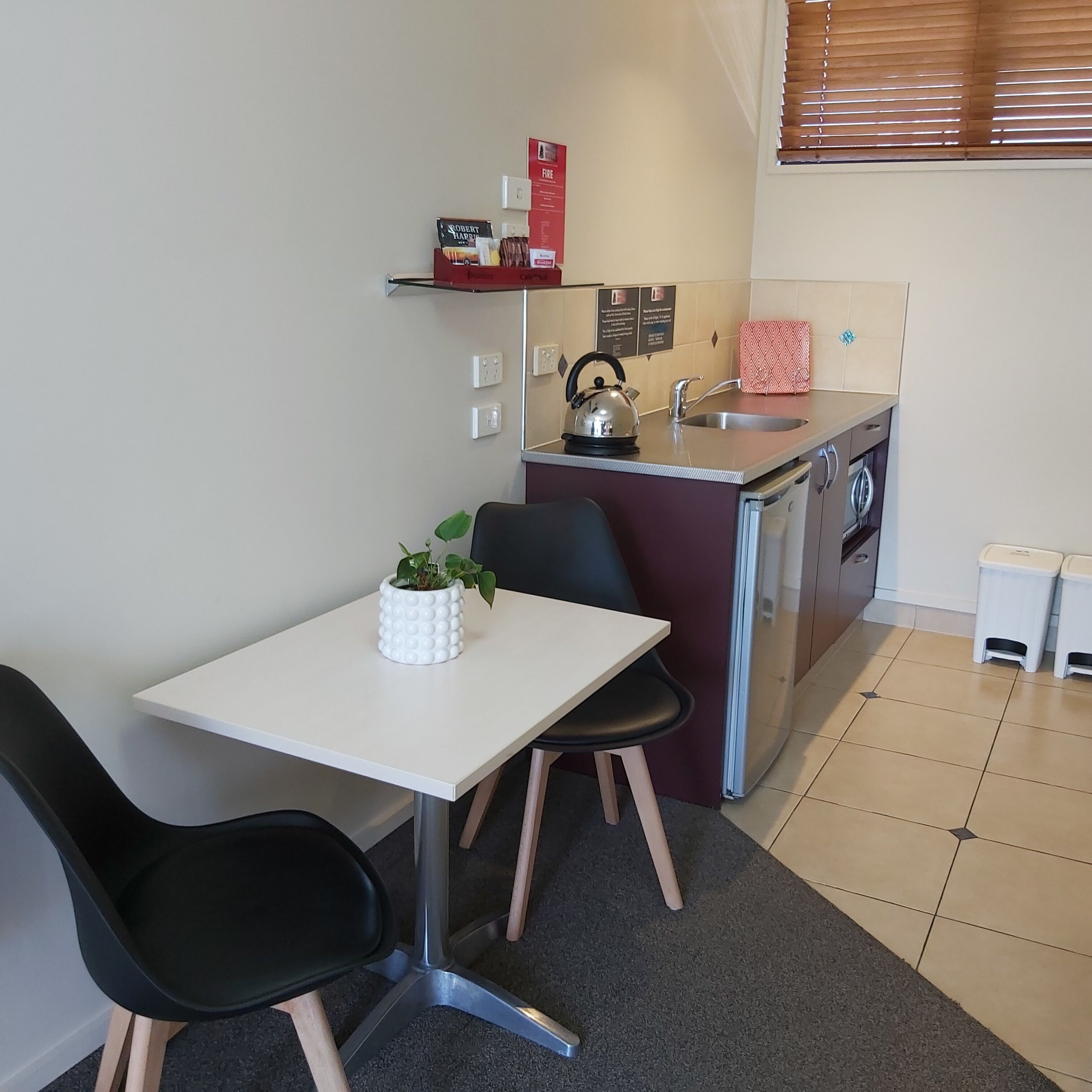 A compact kitchen with a white table, black chairs, a kettle, and a small sink.