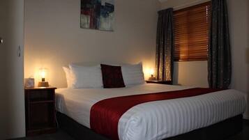 Een net opgemaakt bed met een rode deken, een nachtkastje met een lamp en een raam met gordijnen.
