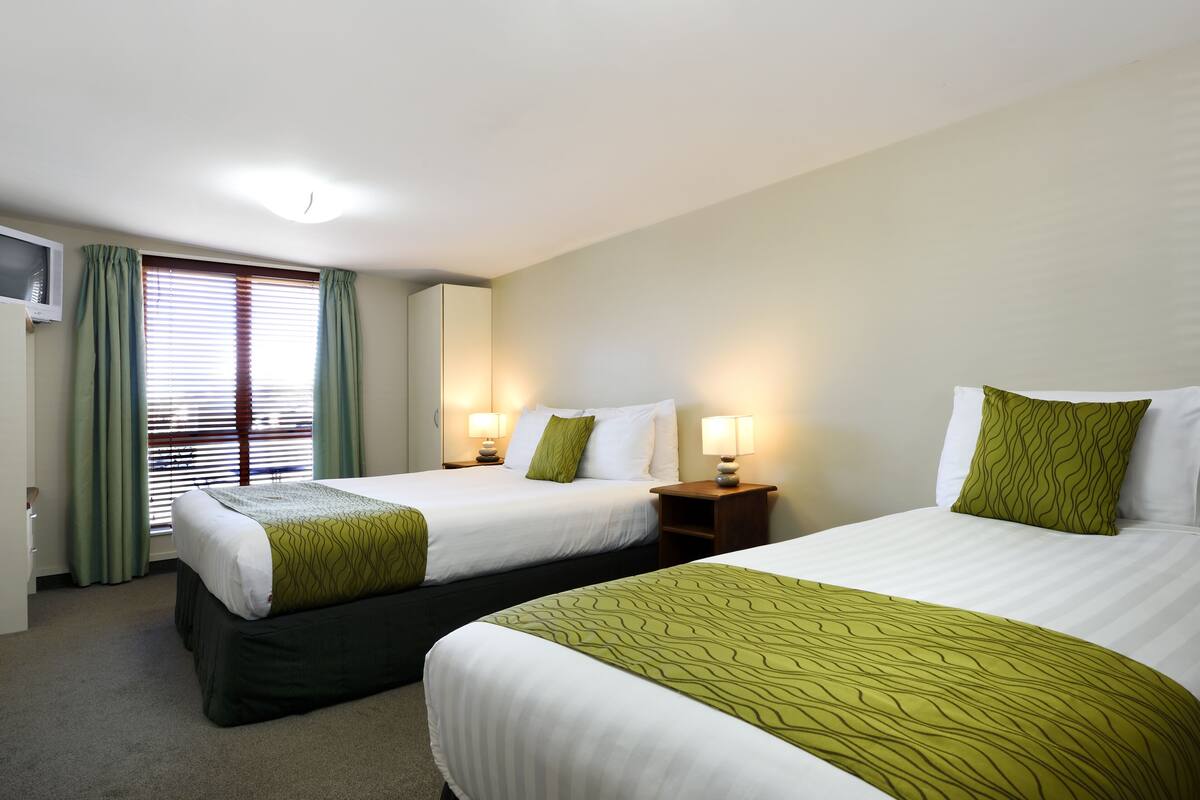 Ett hotellrum med två sängar, grönt och vitt sänglinne, sänglampor, ett fönster med gardiner och en väggmonterad TV.
