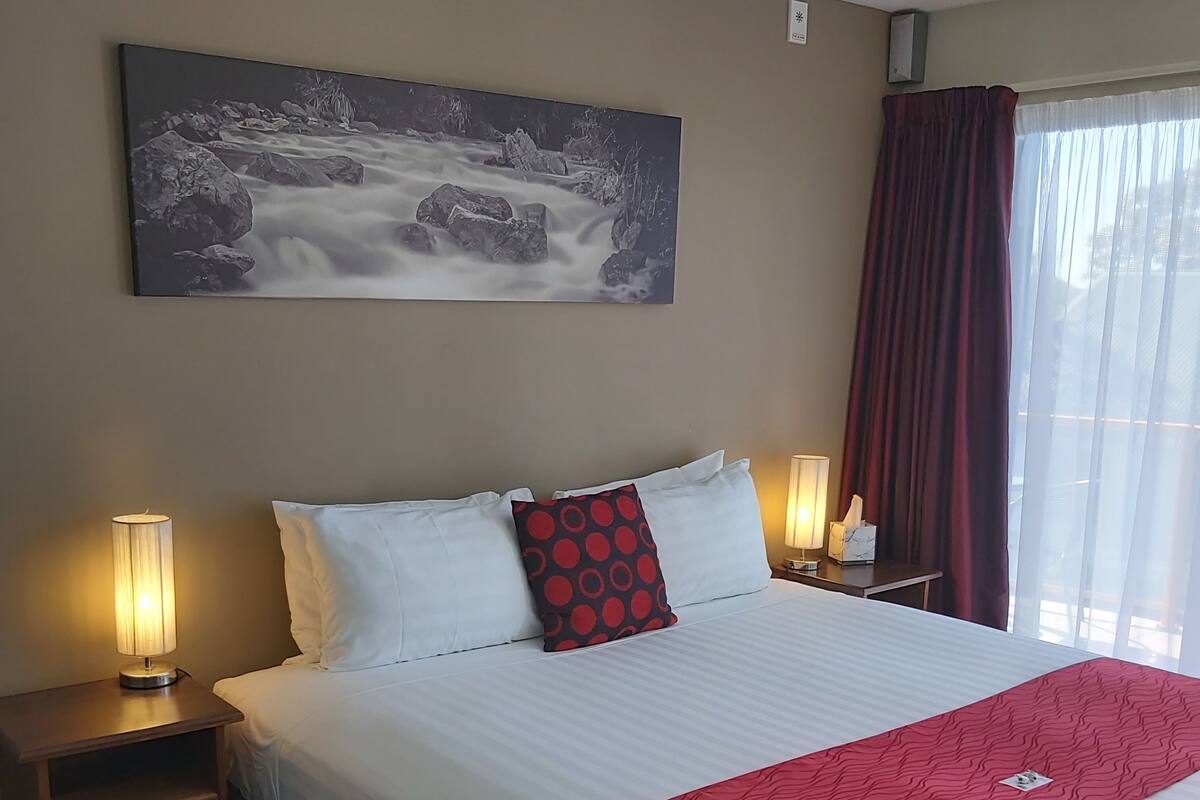 Ett hotellrum med en säng, två sänglampor, en tavla på väggen och ett fönster med gardiner.