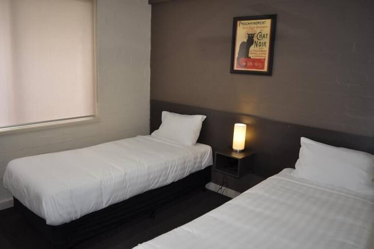 Habitación de hotel con dos camas individuales, una mesita de noche con lámpara y un cartel enmarcados en la pared.