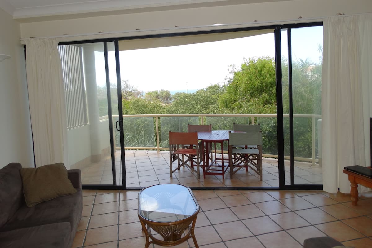 Uma varanda com porta de correr de vidro, uma mesa de jantar com cadeiras, um sofá e uma mesa de centro de vidro.
