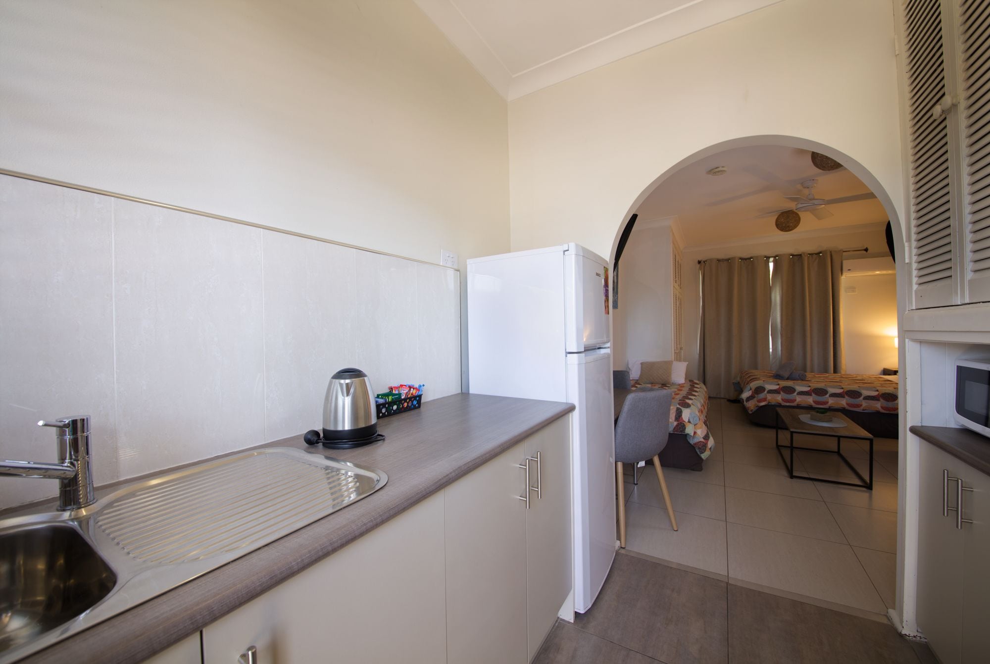 Cocina compacta con lavamanos, hervidor y refrigerador, que da a un pequeño comedor con mesa y sillas.