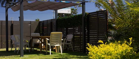 Un patio con mesa y sillas, una pérgola con un letrero y una cerca con follaje al fondo.