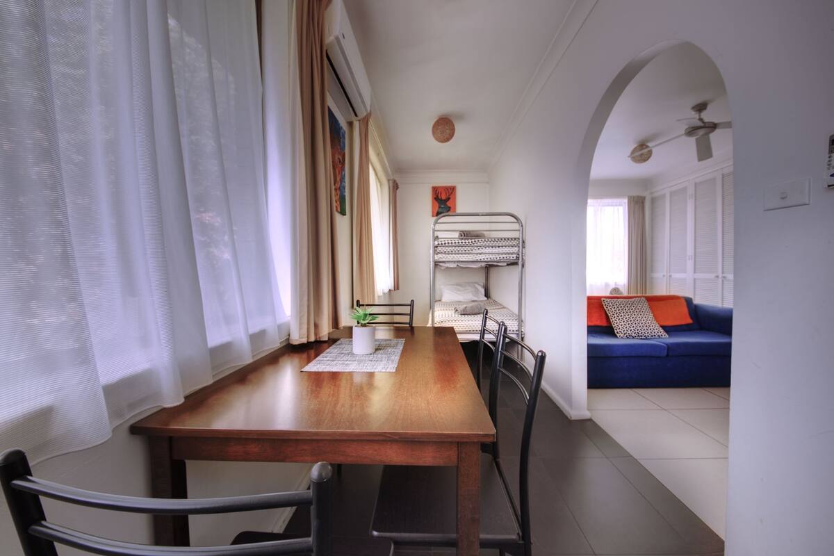 A dining area with a wooden table and chairs, a bunk bed in the background, and a blue sofa with a patterned pillow.