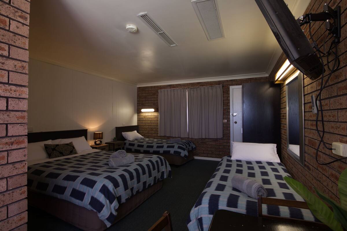 A hotel room with two beds, a brick wall, a window with curtains, a mirror, and a ceiling vent.