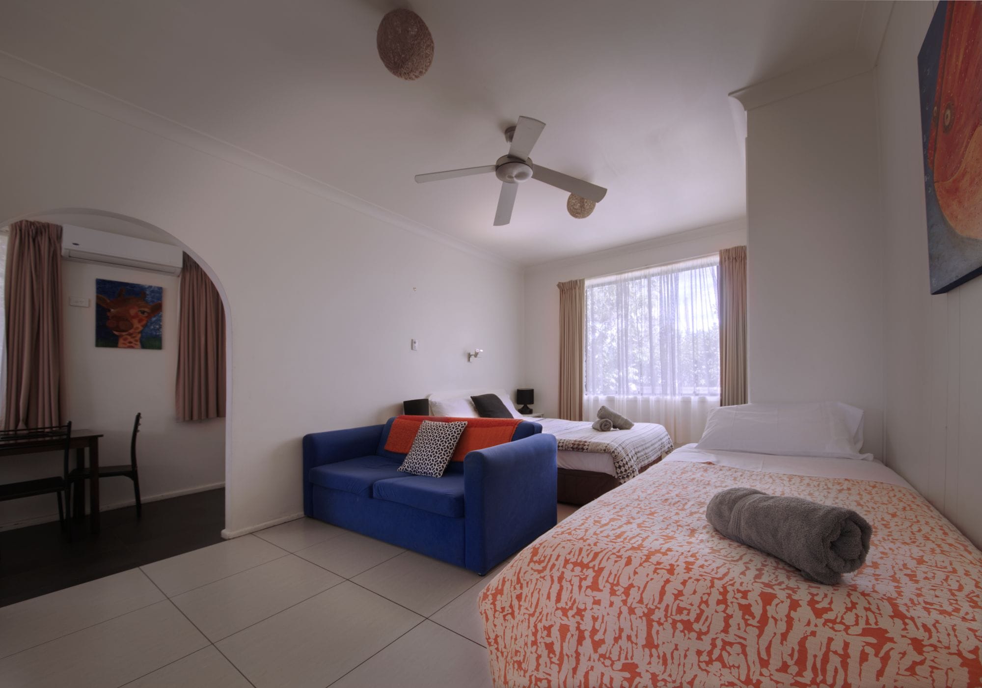A hotel room with a bed, a blue sofa, a ceiling fan, and a window with curtains.