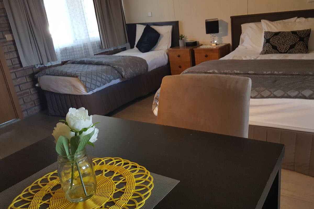 A hotel room with two beds, a dining table with a vase of flowers, and a brick wall.