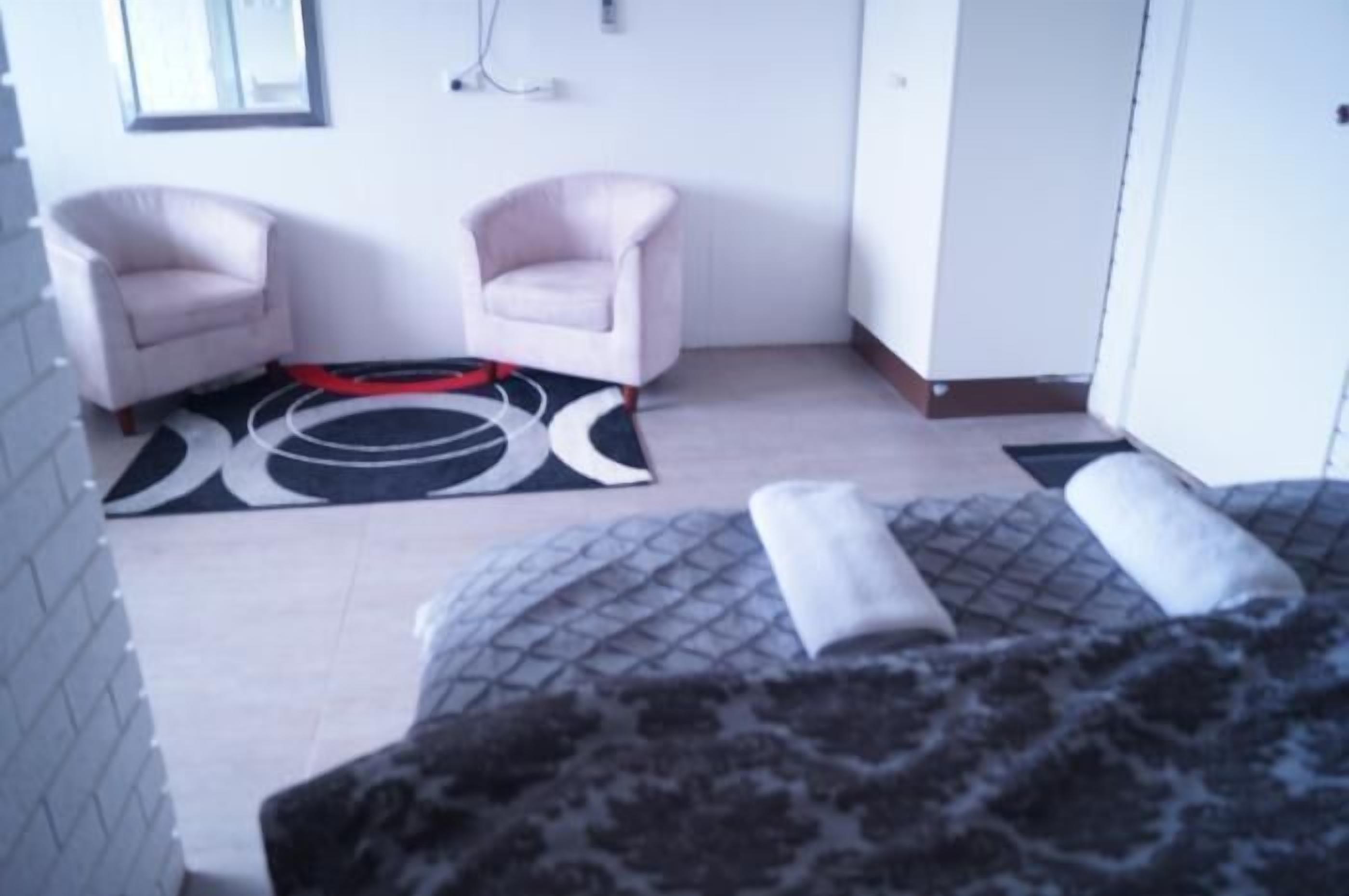 A hotel room with two armchairs, a patterned rug, a bed with a rolled-up blanket, and a tiled floor.
