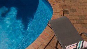 A pool with clear blue water, a brick border, and a lounge chair with striped cushions.