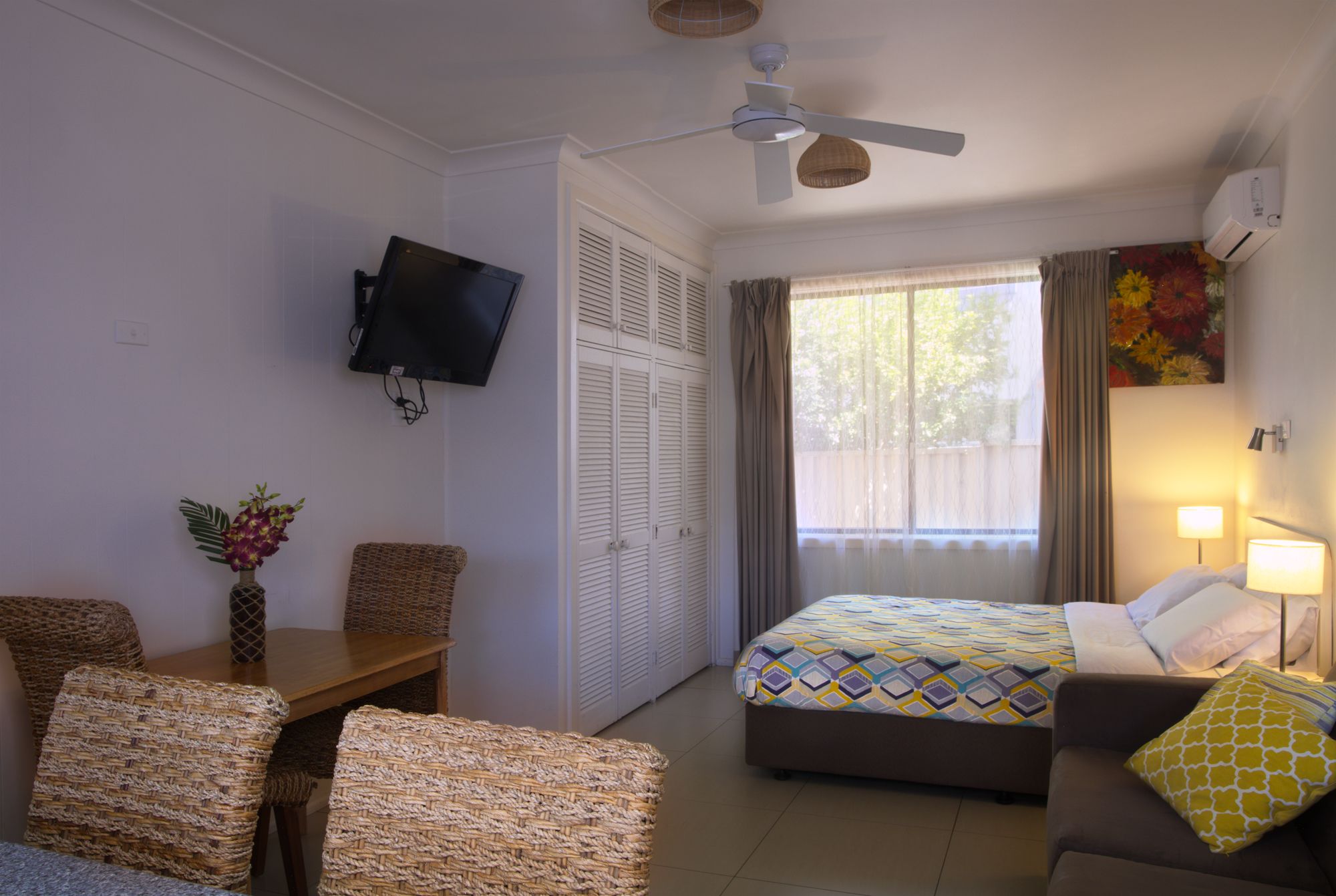 A hotel room with a bed, a sofa, a TV mounted on the wall, a ceiling fan, and a window with curtains.