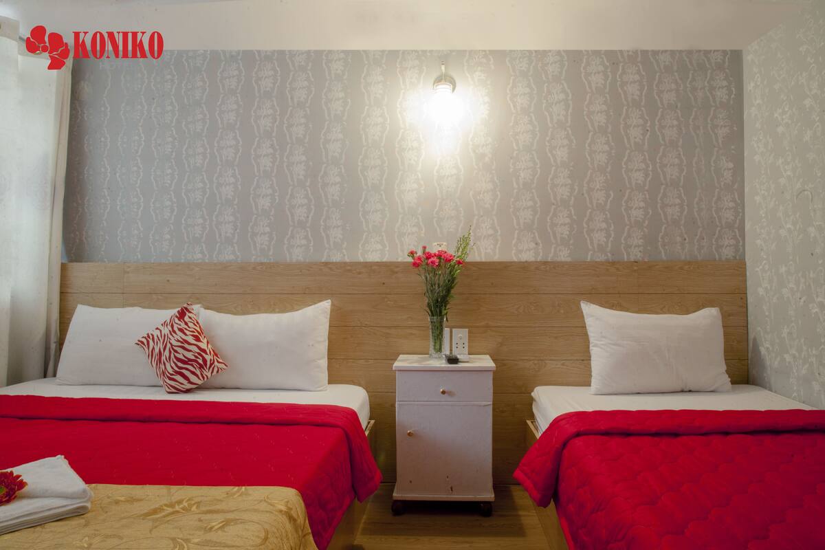Quarto de hotel com duas camas, cada uma com colcha vermelha e almofadas brancas. Entre as camas, um criado-mudo com um vaso de flores e uma luminária. As paredes possuem papel de parede estampado, e há uma cabeceira de madeira.