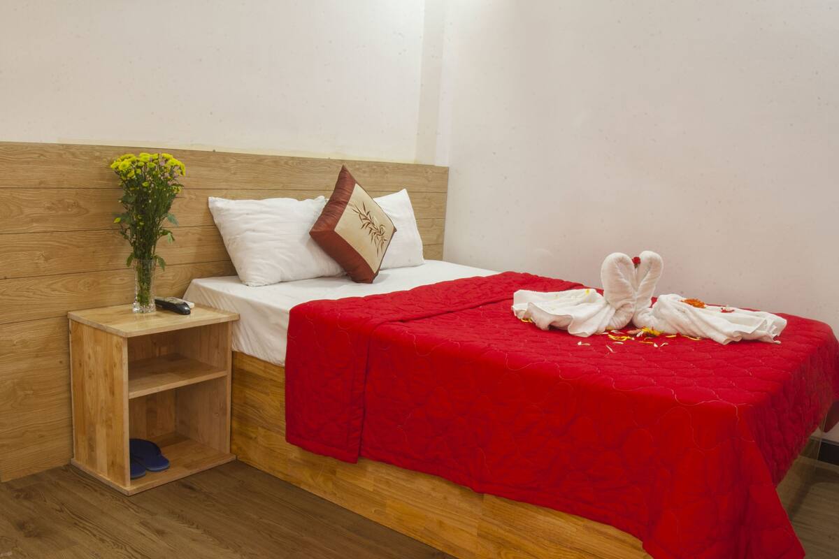 Quarto de hotel com cabeceira de madeira, colcha vermelha, almofada branca, um vaso com flores e um par de toalhas dispostos na cama.