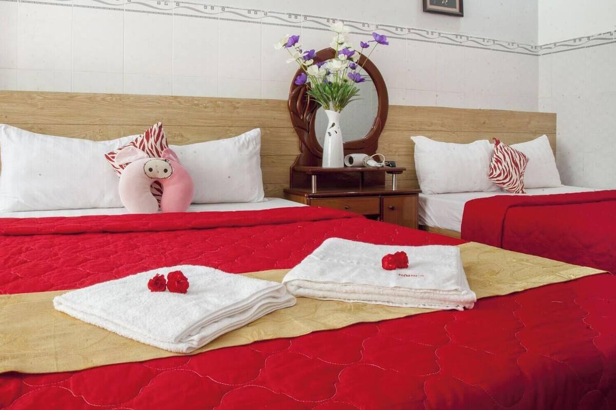 Quarto de hotel com cabeceira de madeira, colcha vermelha, duas almofadas brancas, um brinquedo de pelúcia rosa, um vaso com flores e um espelho na mesa de cabeceira.