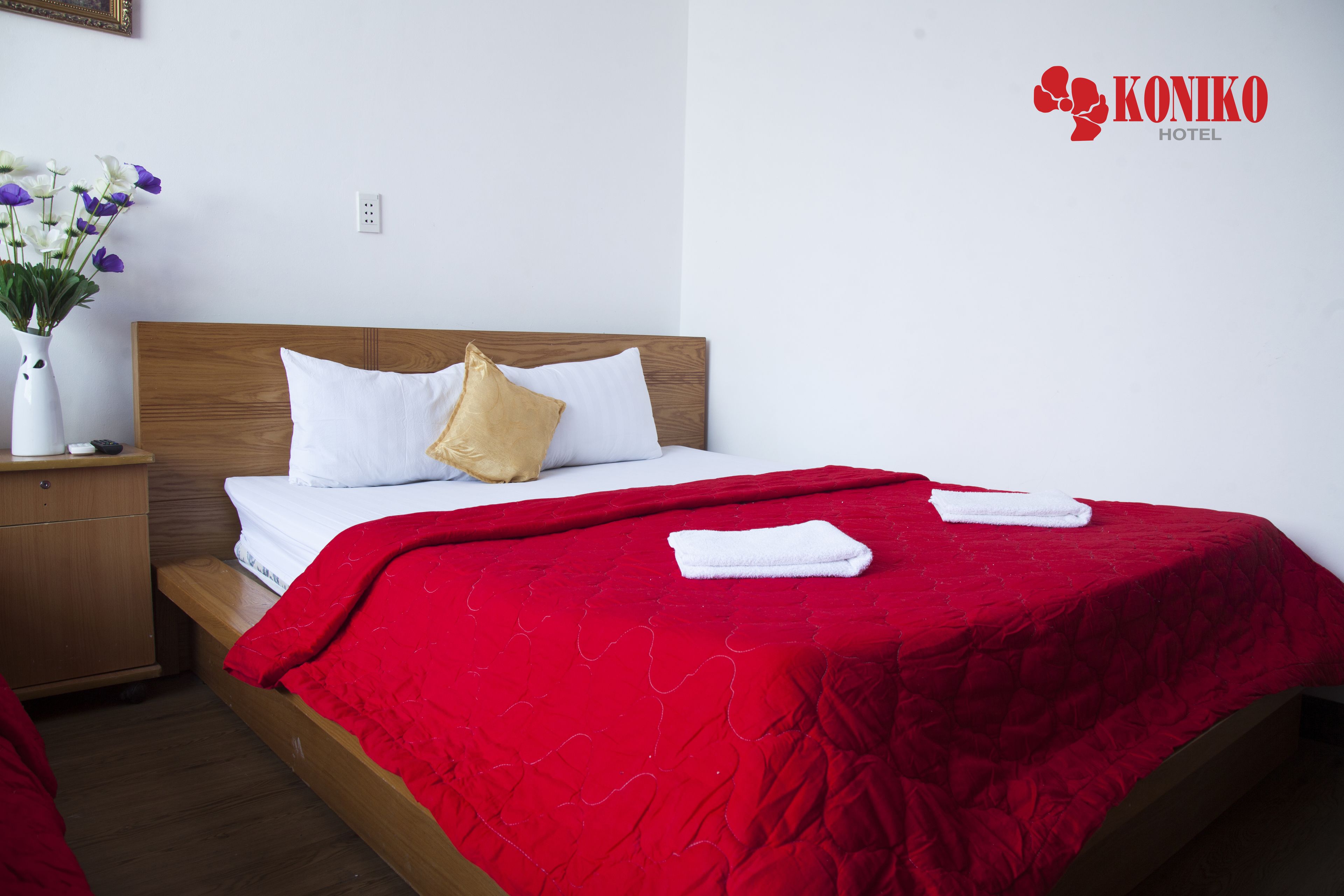 A hotel room with a wooden bed, white bedding, a red blanket, and a vase with flowers on the nightstand.