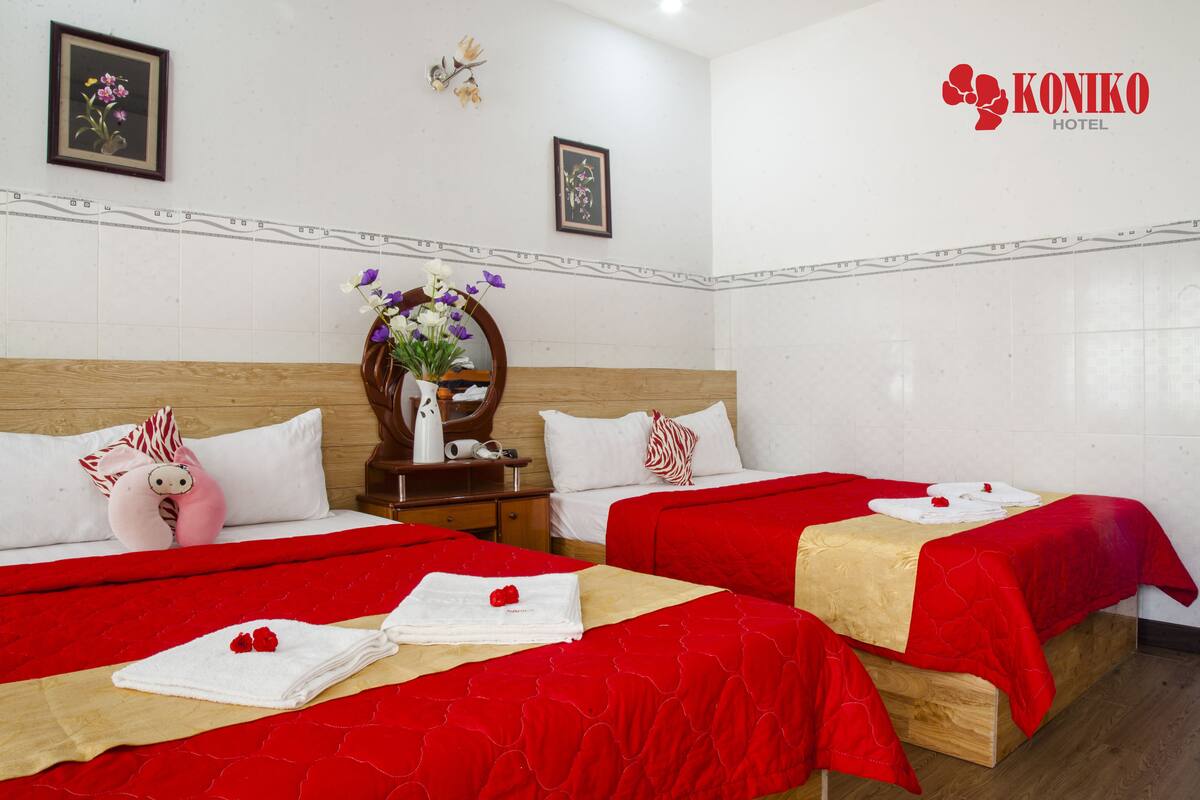 Quarto de hotel com duas camas, cada uma com roupa de cama vermelha e toalhas brancas. Há um vaso com flores na mesa de cabeceira entre as camas.