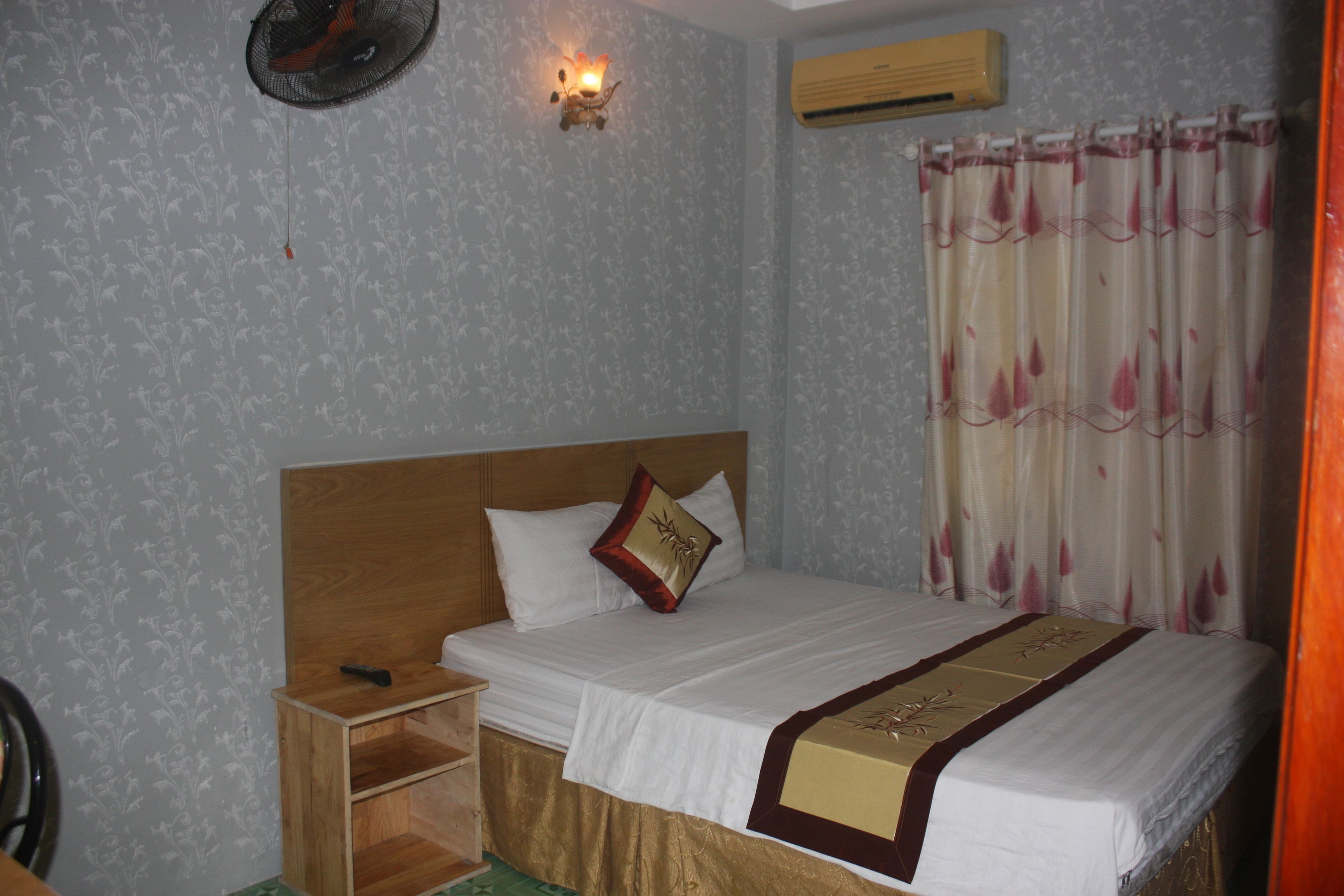 A bedroom with a wooden headboard, a bed with white bedding and a decorative runner, a nightstand with a lamp, and a wall-mounted air conditioner.
