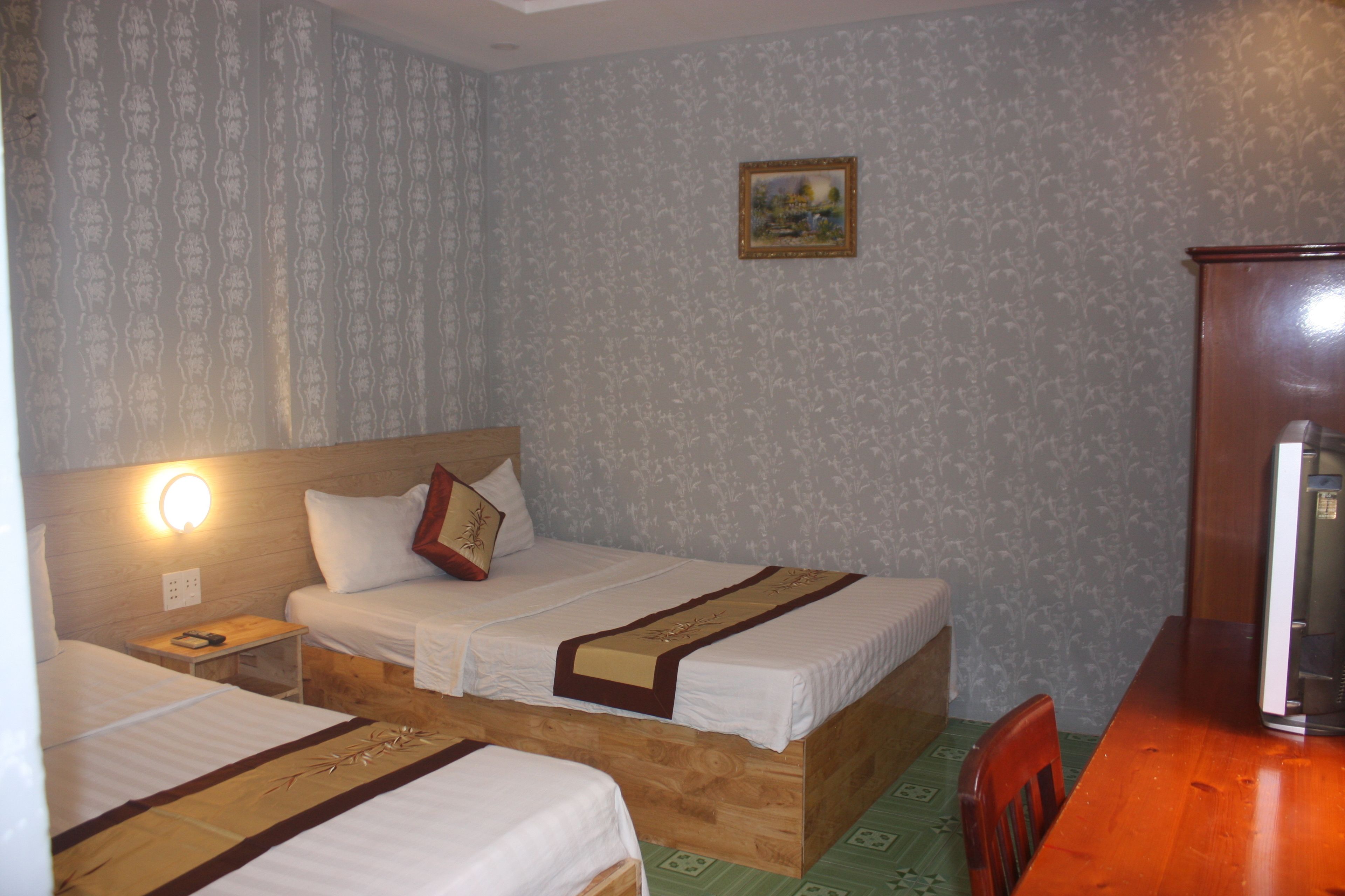 Ein Hotelzimmer mit zwei Einzelbetten, einem hölzernen Kopfteil, einem kleinen Nachttisch mit Lampe, einem gerahmten Bild an der Wand und einem Holztisch mit Stuhl.