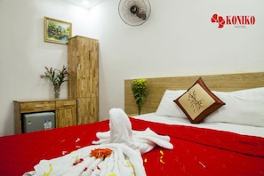 A hotel room with a wooden wardrobe, a small refrigerator, a bed with a red blanket and a white towel, a pillow with a floral design, and a wall-mounted fan.