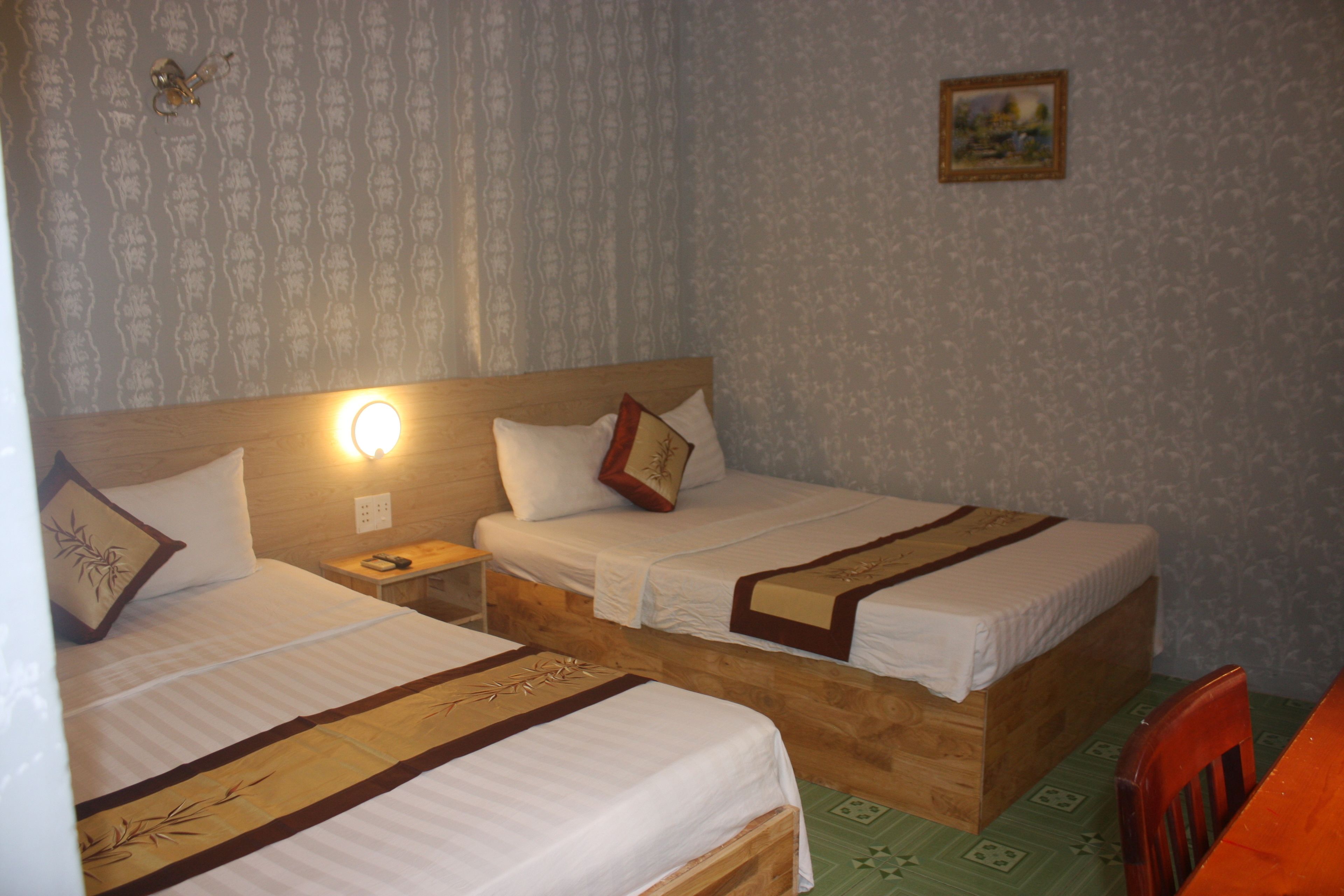 Une chambre avec deux lits en bois, chacun recouvert d’une courtepointe blanche et agrémenté de coussins décoratifs. Une table de chevet avec une lampe se trouve entre les lits, ainsi qu’une petite table avec une chaise. La chambre est décorée de papier peint à motifs et un tableau encadré orne le mur.