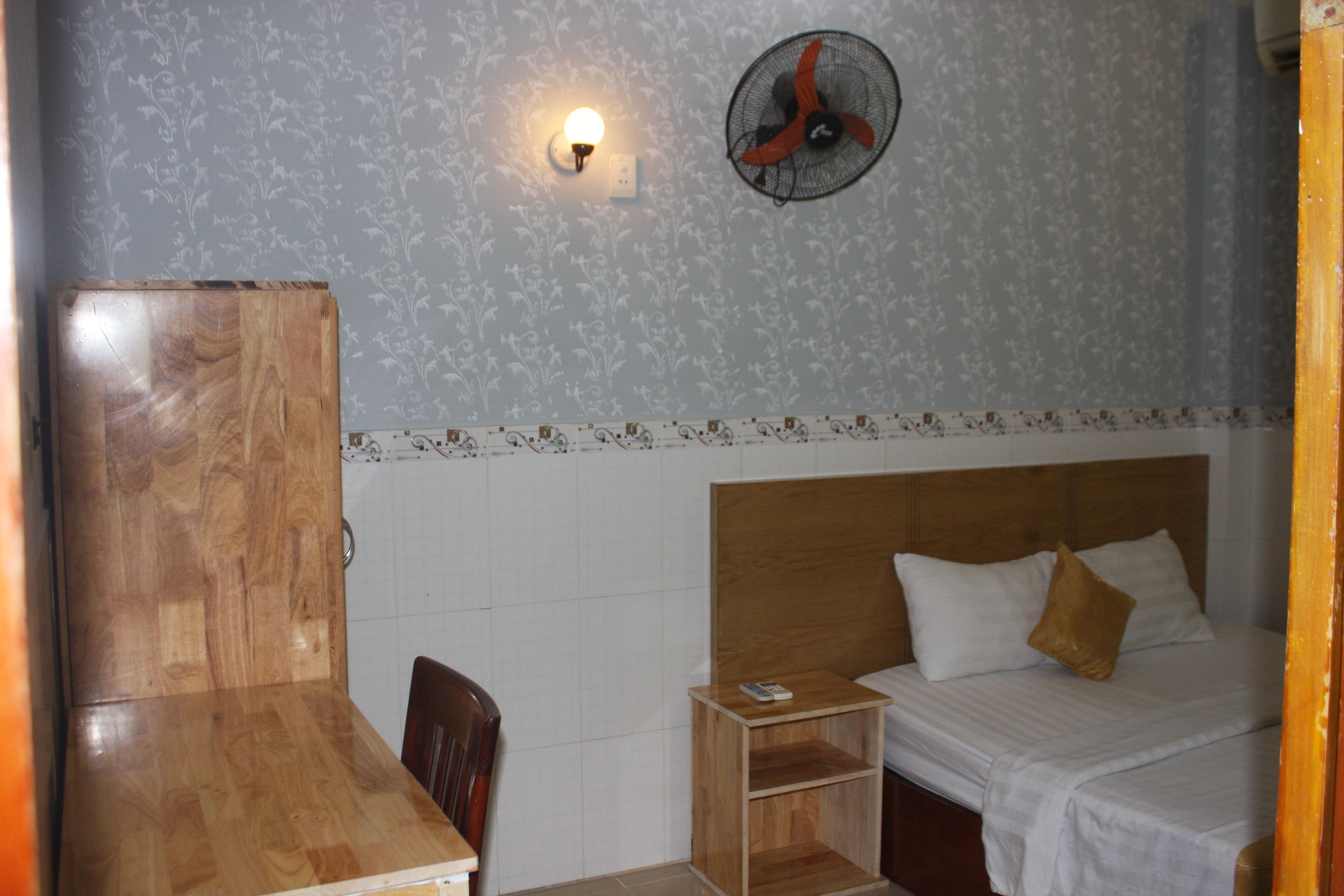 A bedroom with a wooden headboard, a single bed with white bedding, a nightstand, a chair, a wall-mounted fan, and a light fixture.