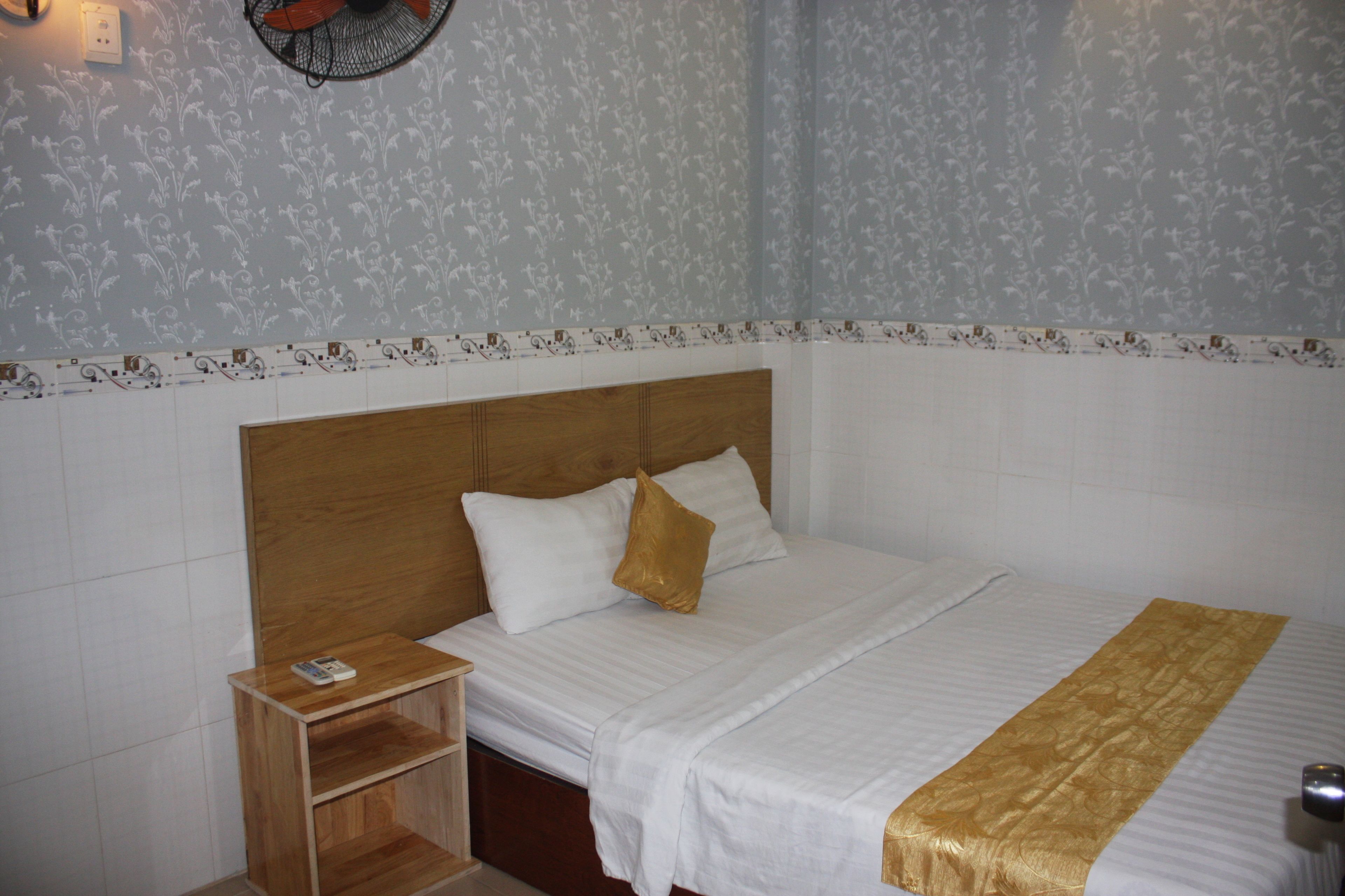 Een net opgemaakt bed met een houten hoofdbord, een nachtkastje met een afstandsbediening, en een behang met een patroon.