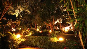 Un jardín nocturno con senderos, plantas iluminadas y una edificación pequeña al fondo.