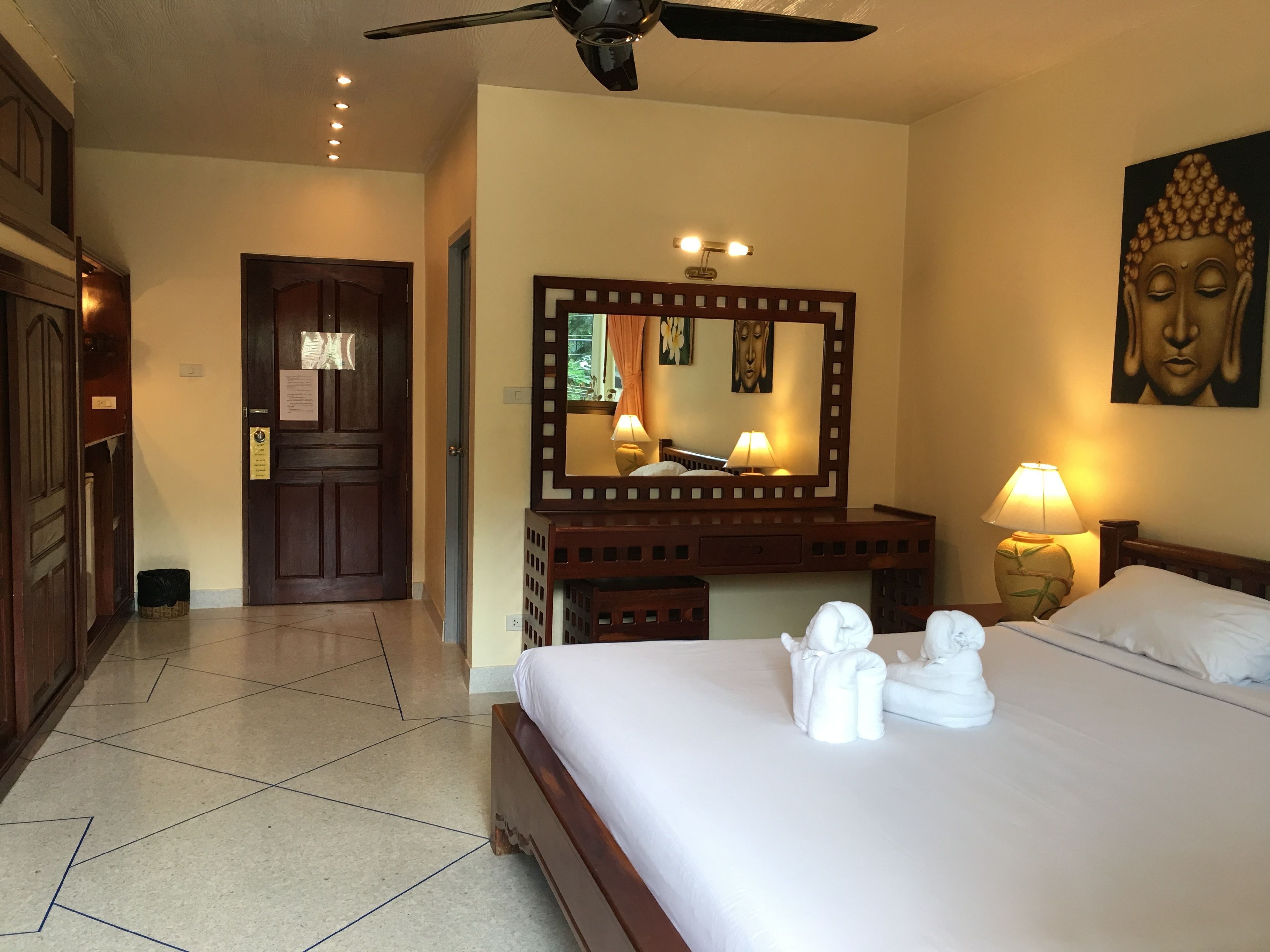 Ein Hotelzimmer mit Bett, einem großen Spiegel, einem Deckenventilator und einem Buddha-Gemälde.